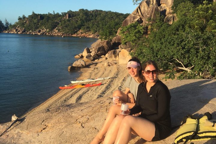 2 girls on shore with drinks and kayak on beach during sunset